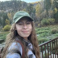 An adult white woman with light brown hair, wearing a flannel and a green cap, stands on a bridge over a pond in a forest.